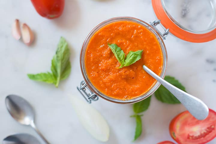 An overhead image of a spoon dipped into a glass jar of Roasted Tomato Sauce made with roma tomatoes, garlic, yellow onion, carrot, olive oil, basil, salt and pepper.